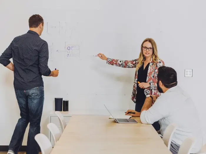 Group of three professionals during a meeting as one woman presents ideas on a whiteboard in an office on Town Service Search